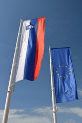 Flaga Słowenii i Unii Europejskiej wiszą obok siebie na drzewcach, powiewają na wietrze, w tle błękitne niebo