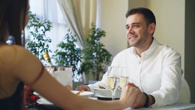 Young handsome man is talking to his girlfriend holding her hand, they are clinking glasses and drinking champagne while having dinner in classy restaurant.