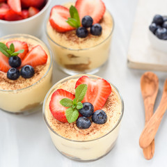homemade, exquisite dessert tiramisu in glasses decorated with strawberry, blueberry, mint on white wooden table, top view.