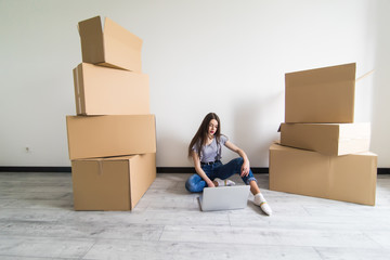 Young woman sitting on hardwood floor in new aprtmanet, using laptop next to cardboard box