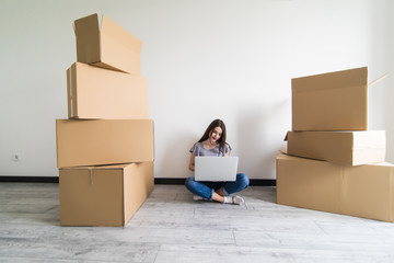 Happy young woman sitting on floor near moving boxes. Young woman moving to new home. Woman using laptop, looking at camera and smiling