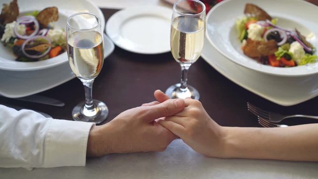 Close-up shot of male hand holding and caressing female hand on table with champagne glasses and plates. Romantic relationship, love and fine dining concept.