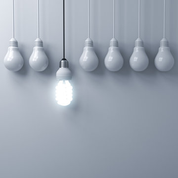 One hanging eco energy saving light bulb glowing and standing out from unlit incandescent bulbs on white wall background with shadow , leadership and different creative idea concepts . 3D rendering.