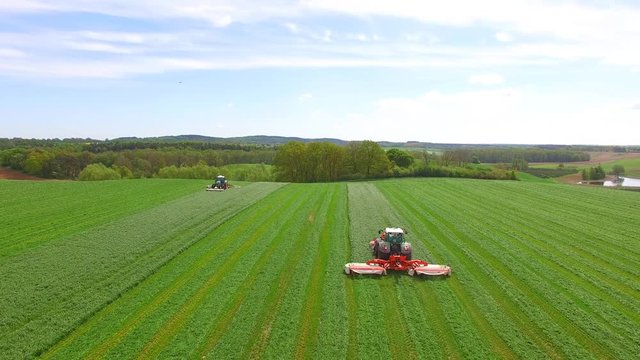 Aerial view of two modern tractors mowing a green fresh grass field on a sunny day with blue sky. 