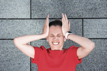 A young woman in a red business dress is standing opposite the granite wall, holding both hands behind her head because of a very strong headache.