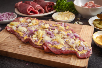 preparing beef roulades, fresh raw meat coated with mustard and onions on a wooden cutting board, ingredients as pickled cucumber and bacon, dark kitchen countertop