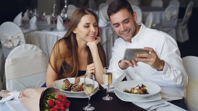 Happy attractive young people are watching smartphone together, smiling and talking while having dinner in restaurant. Modern technologies and romance concept.