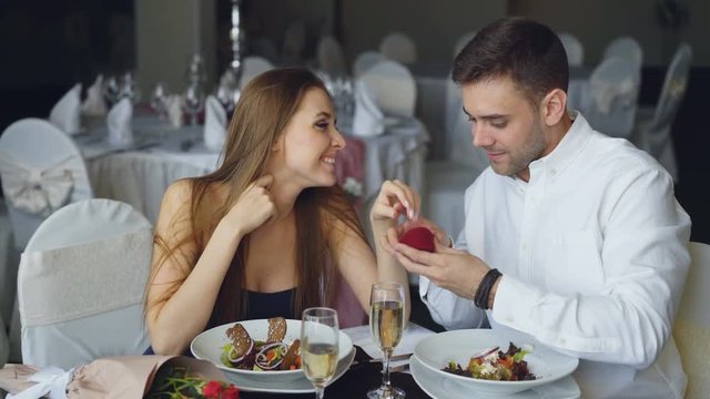 Attractive woman is saying yes to marriage proposal and her boyfriend is putting engagement ring on her finger and kissing her. Romantic relationship and restaurant date concept.