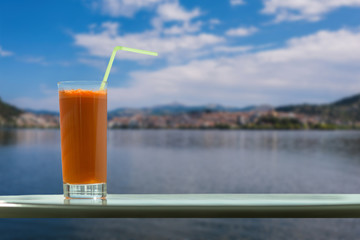 A glass of carrot juice with a straw in the cafe on the background of Kastoria town and Orestias lake. Greece