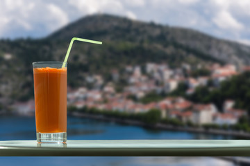 A glass of carrot juice with a straw in the cafe on the background of Kastoria town and Orestias lake. Greece