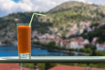 A glass of carrot juice with a straw in the cafe on the background of Kastoria town and Orestias lake. Greece