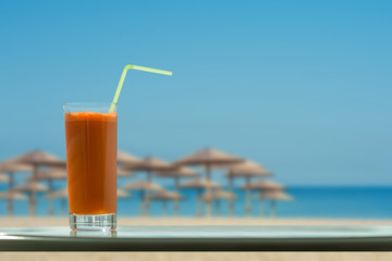 A glass of carrot juice with a straw in the cafe on the beach and straw umbrellas backgrounds 