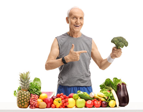 Elderly man behind a table with fruit and vegetables holding broccoli and pointing