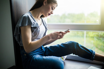 Fototapeta na wymiar Happy woman in t-shirt sitting on the windowsill and writing message on smartphone at home