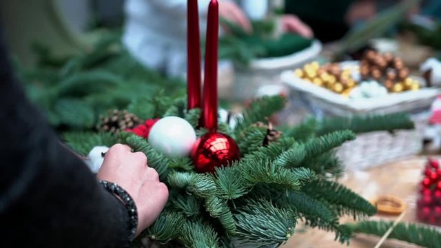 Female hands attach cotton ball flower to handcrafted table centerpiece decoration: basket with fir-tree branches, pinecones, christmas tree balls and taper candles on plastic wrap, close-up. 