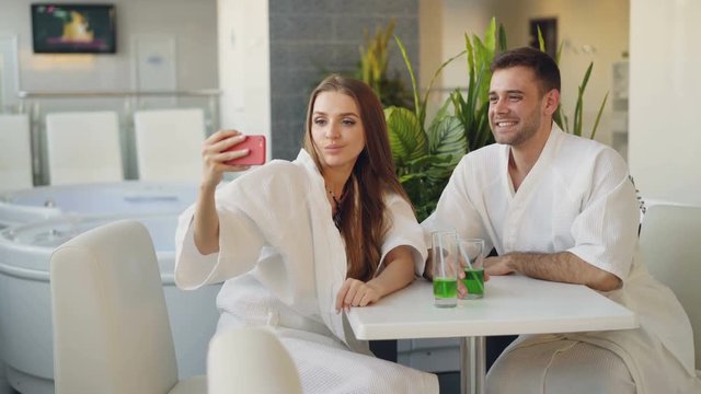 Pretty girl and her loving boyfriend are taking selfie with cocktail glasses using smartphone while relaxing in spa salon. They are smiling and posing looking at camera.
