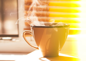 The big ceramics cup of strong coffee on a window sill in a room in warm sunbeam of golden light.Aroma steam above the cup. Traditional breakfast detail.Hot fresh drink. Good morning concept.