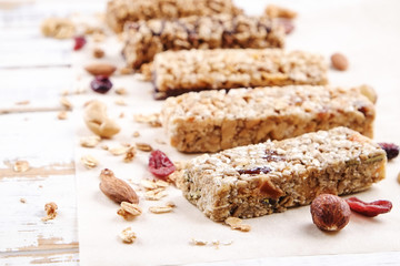 Various granola energy bars in row with scattered mixed nuts, cereals & dried fruit, grunged white wood table background. Healthy nutritious vegan fitness food snack. Top view, copy space, close up.