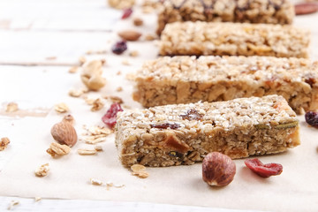 Various granola energy bars in row with scattered mixed nuts, cereals & dried fruit, grunged white wood table background. Healthy nutritious vegan fitness food snack. Top view, copy space, close up.