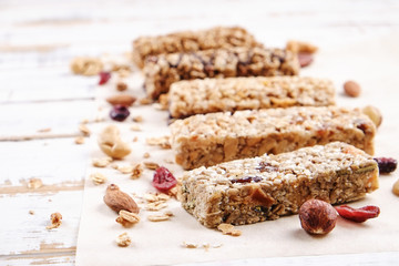 Various granola energy bars in row with scattered mixed nuts, cereals & dried fruit, grunged white wood table background. Healthy nutritious vegan fitness food snack. Top view, copy space, close up.