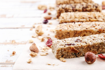 Various granola energy bars in row with scattered mixed nuts, cereals & dried fruit, grunged white wood table background. Healthy nutritious vegan fitness food snack. Top view, copy space, close up.
