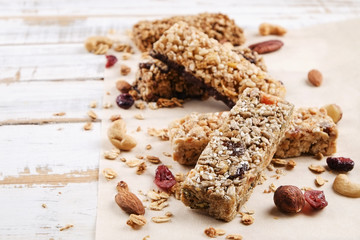 Various granola chocolate energy bars in row with scattered mixed nuts, cereals & dried fruit, grunged white wood table background. Healthy vegan fitness food snack. Top view, copy space, close up.