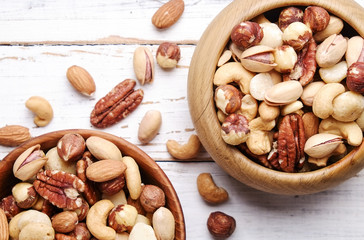 Mixed nuts in wooden bowl and scattered on table. Trail mix of pecan, almond, macadamia & brazil edible nuts with walnut hazelnut on wood textured surface. Background, copy space, top view, close up.