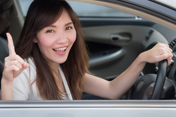 Fototapeta na wymiar smiling woman driver driving and pointing up one finger; portrait of happy, joyful asian woman driver in left hand driving style pointing up number one gesture; 30s adult asian Chinese woman model