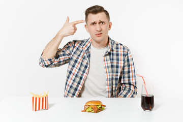 Man point fingers to head as if he shoot himself, sits at table with burger french fries cola in glass isolated on white background. Proper nutrition or American classic fast food. Area to copy space.