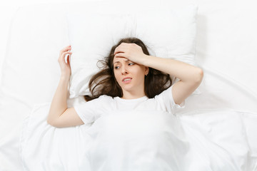 Top view tired brunette young woman looking aside, lying in bed with white sheet, pillow, blanket. Shocked female put hand on head with headache, spending time in room. Rest, relax, good mood concept.