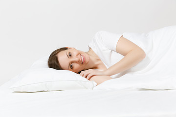 Obraz na płótnie Canvas Calm young brunette woman lying in bed with white sheet, pillow, blanket on white background. Smiling beauty female spending time in room. Rest, relax, good mood concept. Copy space for advertisement.