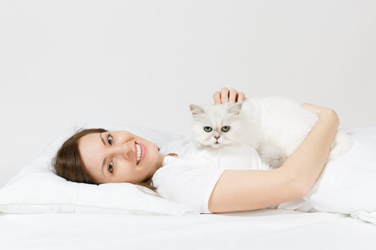 Calm young woman lying in bed with white cute Persian silver chinchilla cat, sheet, pillow, blanket on white background. Beauty female spending time in room. Rest, relax, good mood concept. Copy space