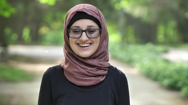 Portrait of a young smiling girl in glasses wearing hijab, outdoor, in a park in the background. 50 fps