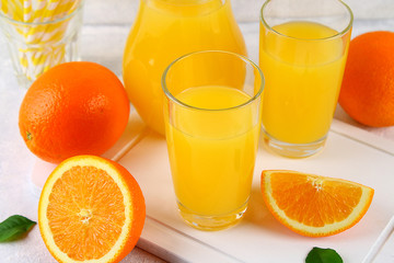 Glass cups and a pitcher of fresh orange juice with slices of orange and yellow tubes on a light gray table.