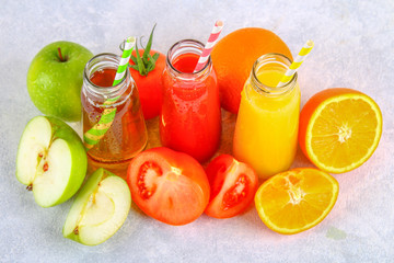 Bottles with fresh orange, apple, tomato juice and colored tubules on a gray concrete table.
