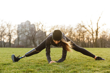 Photo of handicapped sportswoman in black tracksuit, stretching prosthesis leg and sitting on twine...