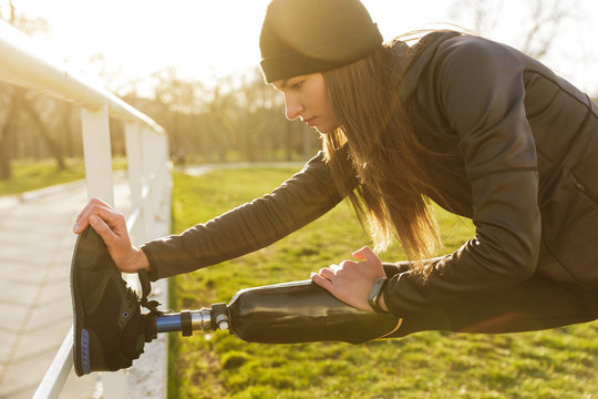 Image of disabled running woman in sportswear, doing slopes and stretching prosthetic leg on grass using railing