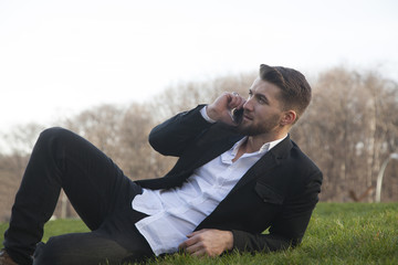 Businessman is lying on a meadow in a city-park and talks on a smart phone