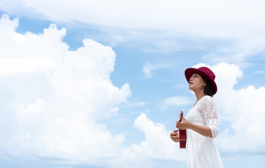 Photo of left side women wearing white dress, red hat playing ukulele outdoor with blue sky and white clouds background. Travel and summer concept.
