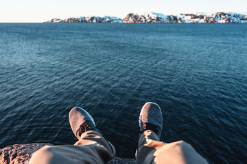 Fototapeta premium Traveler sitting on cliff in front of breakage above ocean and snow mountains. POV view of legs and shoes on background blue water and rock