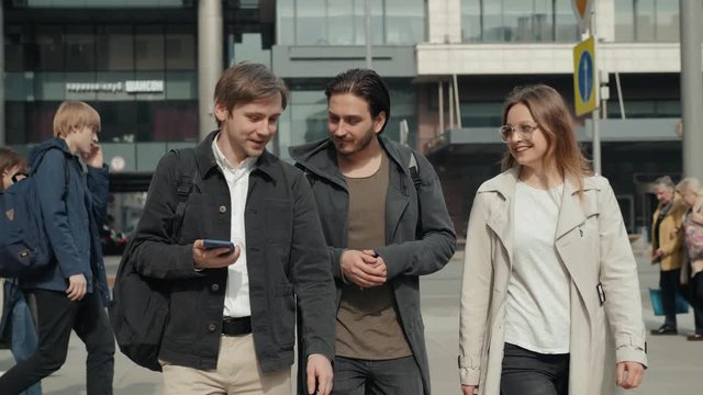 Three amazed students checking on line content on a smart phone in the street, happy friends on city street walking after lessons in university school