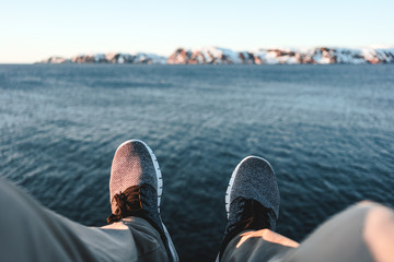 Fototapeta premium Happy traveler sitting on rock and looks on legs and shoes. POV view on feet on background of ocean and mountains. Adventure vacations outdoor concept
