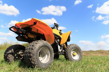 A four-wheeled yellow ATV quad-bike standing idle on the green grass, with trees and a blue sky with clouds on the background