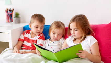 childhood, leisure and family concept - little kids reading book in bed at home