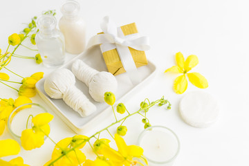 Fototapeta na wymiar Bright spa background: candles, thai massage herbal bags, shampoo bottles, yellow flowers and gift box with ribbon on white. Health, skin treatment, holiday sale concept. Text space