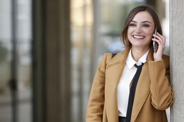 Manager vestita con cappotto beige camicia  bianca e cravatta nera , parla divertita al telefono fuori dal suo uffico