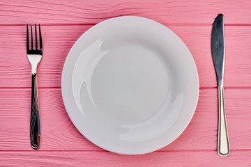 Plate, fork and knife on table. Pink textured wood with white plate, silver fork and knife, top view.
