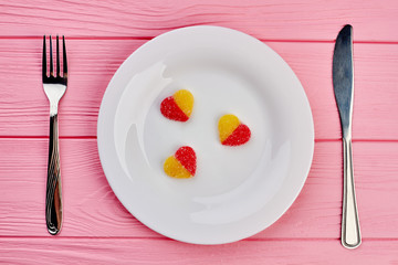 White plate with heart shaped candies. Plate with three jelly hearts, fork and knife on pink wooden background. Valentines Day concept.