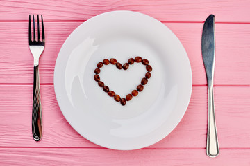 White plate with heart made of coffee beans. White plate with heart shaped coffee beans, fork and knife on pink wooden background, top view.