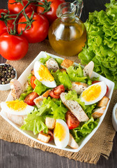 Fresh salad made of tomato, ruccola, chicken breast, eggs, arugula, crackers and spices. Caesar salad in a white, transparent bowl on wooden background